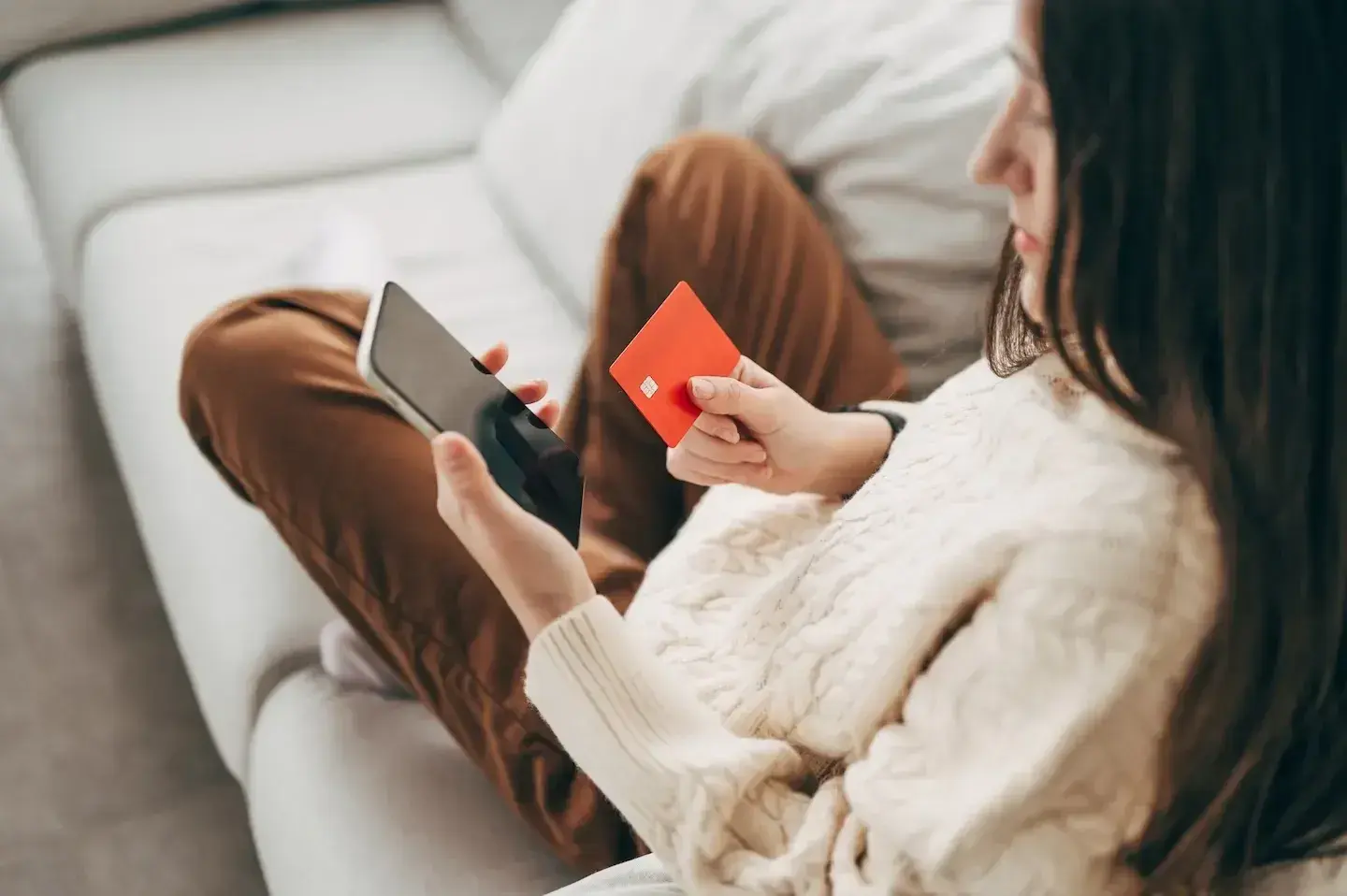 Woman sitting on a couch holding a smartphone and a red credit card.