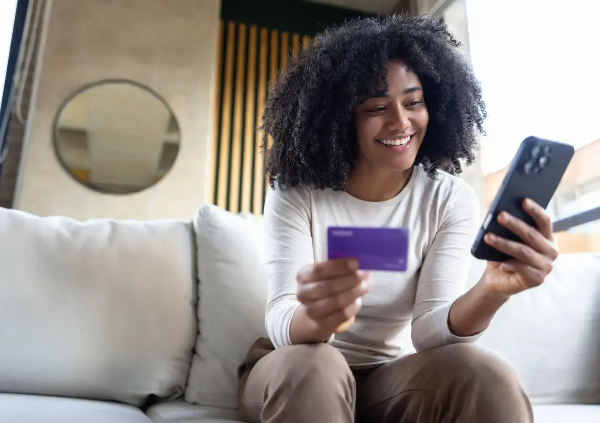 Smiling woman sitting on a couch, holding a purple credit card and smartphone.