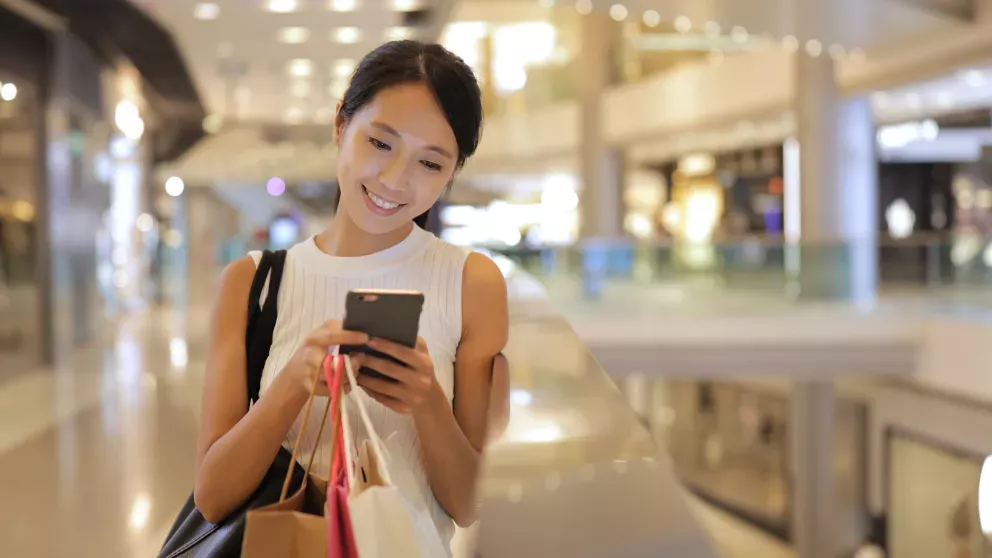 Mulher sorridente no shopping com sacolas de compras olhando para o celular