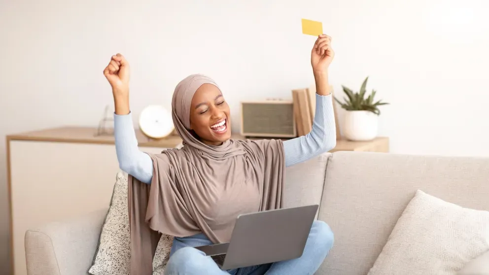 Mulher muçulmana animada segurando cartão e laptop Mulher muçulmana animada segurando cartão e laptop
