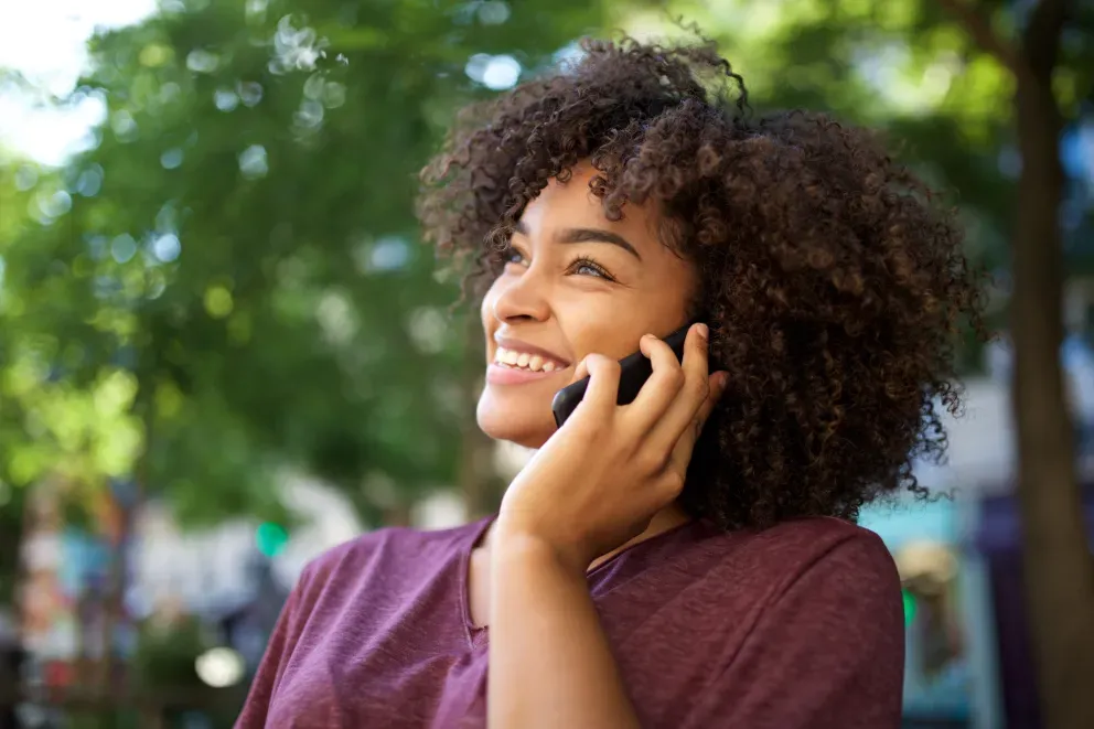 Mulher nova com cabelo cacheado sorrindo enquanto fala ao telefone ao ar livre