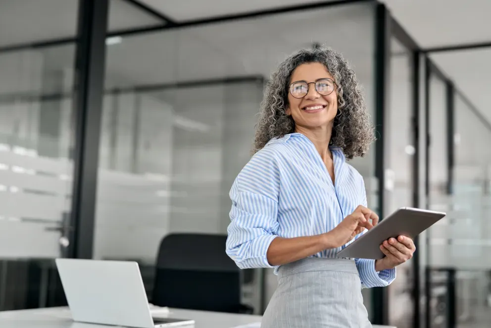 Professional woman with curly hair smiling while holding tablet in modern office with glass walls