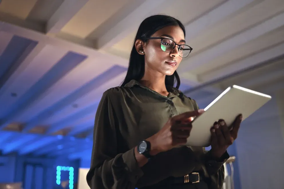 Jovem mulher de óculos segurando um tablet, olhando para cima em um ambiente de escritório moderno mal iluminado.