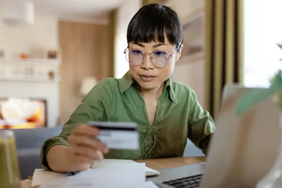 Jovem mulher asiática de camisa verde e óculos segurando um cartão de crédito enquanto faz compras on-line em casa.