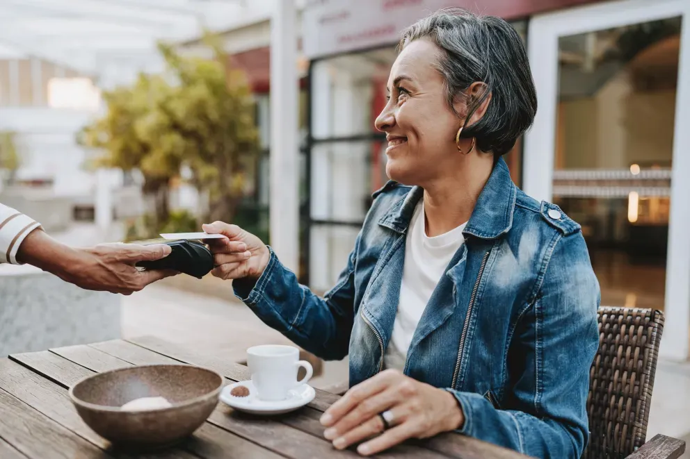 Mulher mais velha sorridente tocando seu telefone em um leitor de cartões em uma mesa de café ao ar livre.