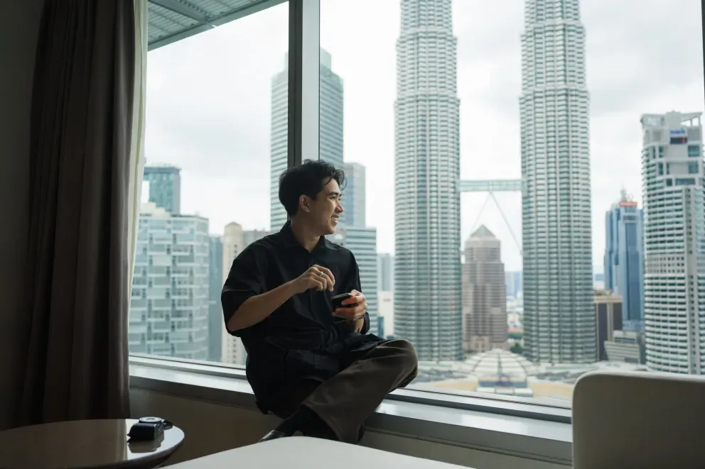 Homem sentado no parapeito de uma janela segurando um copo, com as Torres Petronas de Kuala Lumpur visíveis atrás dele.