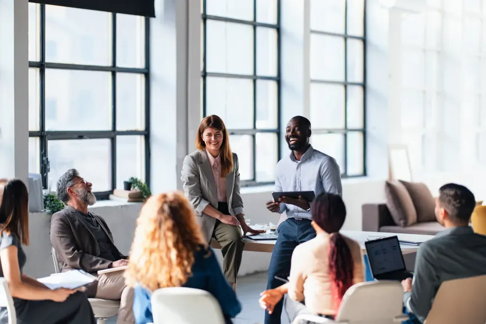 Two professionals leading a group discussion in a bright, modern meeting room with diverse attendees.