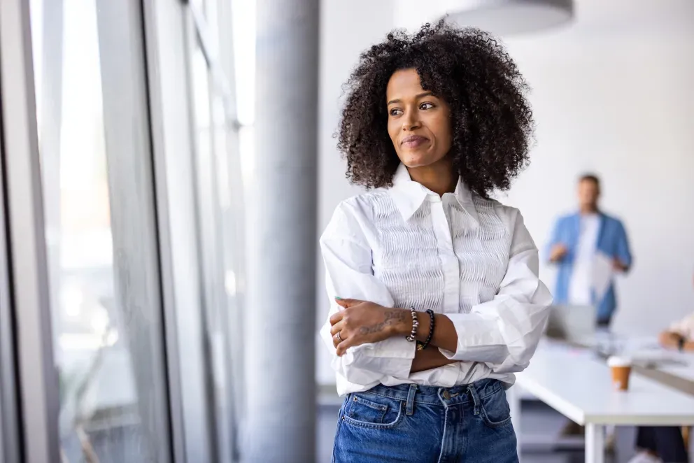 Young Black woman in white blouse and jeans, arms crossed, looking confidently in a bright office.