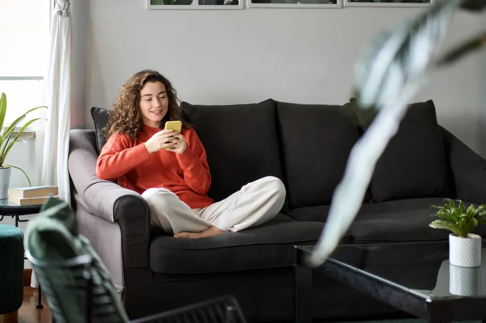 Mulher de suéter vermelho relaxando no sofá escuro, segurando uma bebida em uma sala de estar aconchegante.