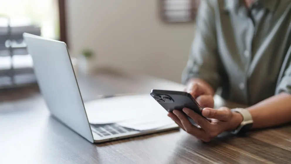 Person using a smartphone at a wooden desk with an open laptop nearby.