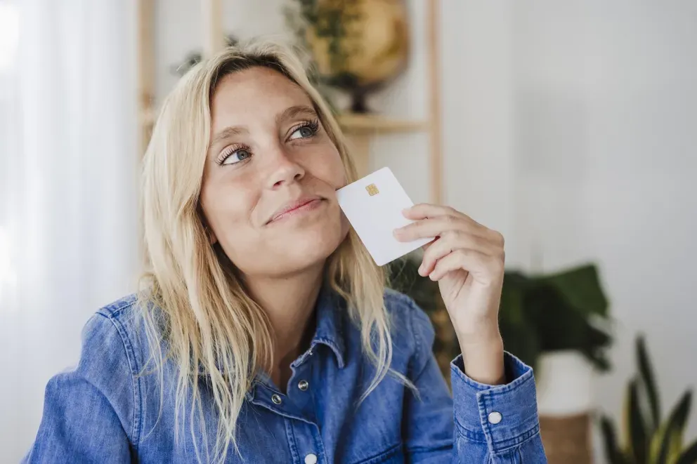 Thoughtful woman in denim shirt holding a small white card, looking upward. Thoughtful woman in denim shirt holding a small white card, looking upward.
