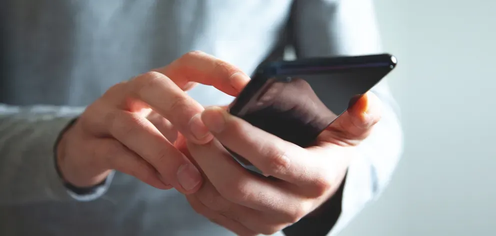 Close-up photo of a person in a gray sweater holding a black smartphone and tapping the screen with one finger.