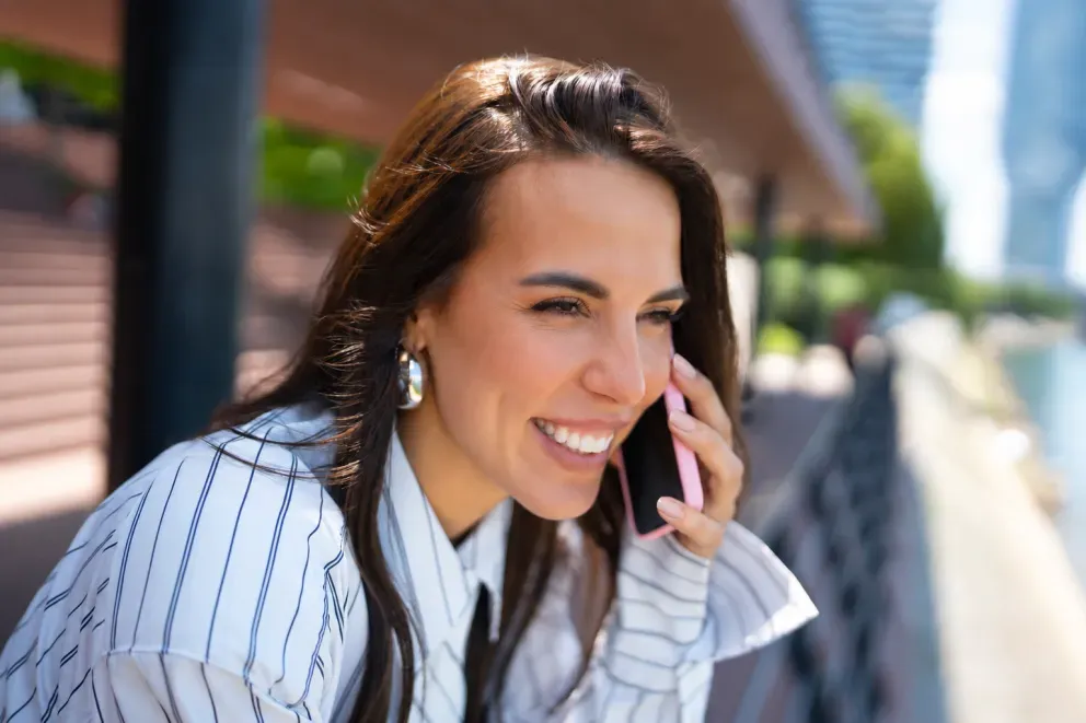 Smiling young woman with dark hair talking on a phone outdoors, wearing a striped blazer. Smiling young woman with dark hair talking on a phone outdoors, wearing a striped blazer.