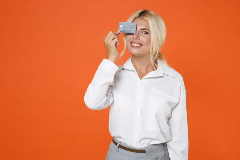 Smiling blonde woman in a white blouse holds a credit card over one eye against a bright orange background.