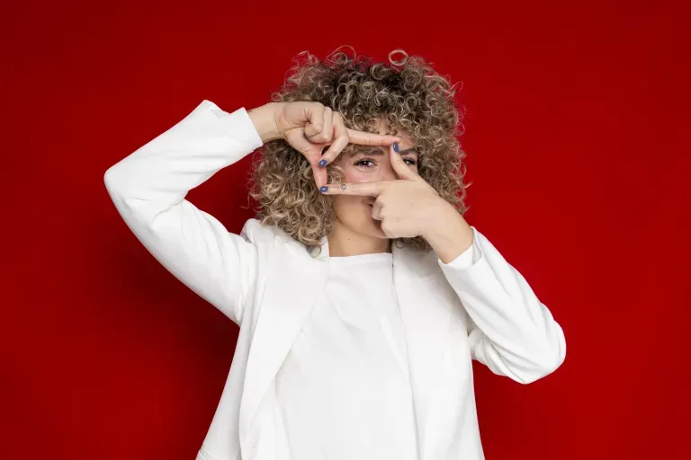 Person with curly blonde hair in a white outfit framing one eye with their fingers against a red background.