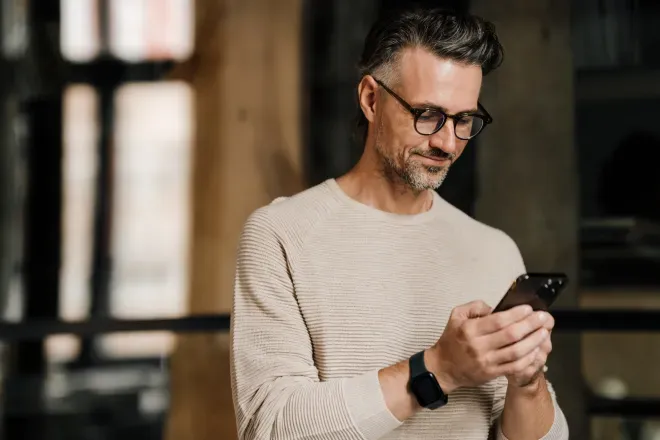 Homem de meia-idade com óculos sorrindo enquanto usa um smartphone dentro de casa.