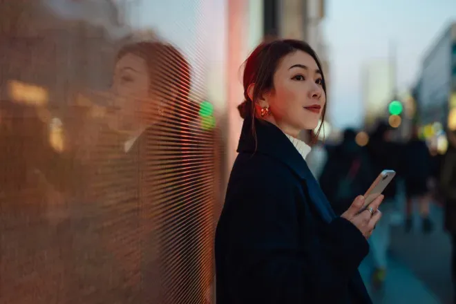 Mulher de casaco escuro segurando smartphone, encostada na parede iluminada ao entardecer. Mulher de casaco escuro segurando smartphone, encostada na parede iluminada ao entardecer.