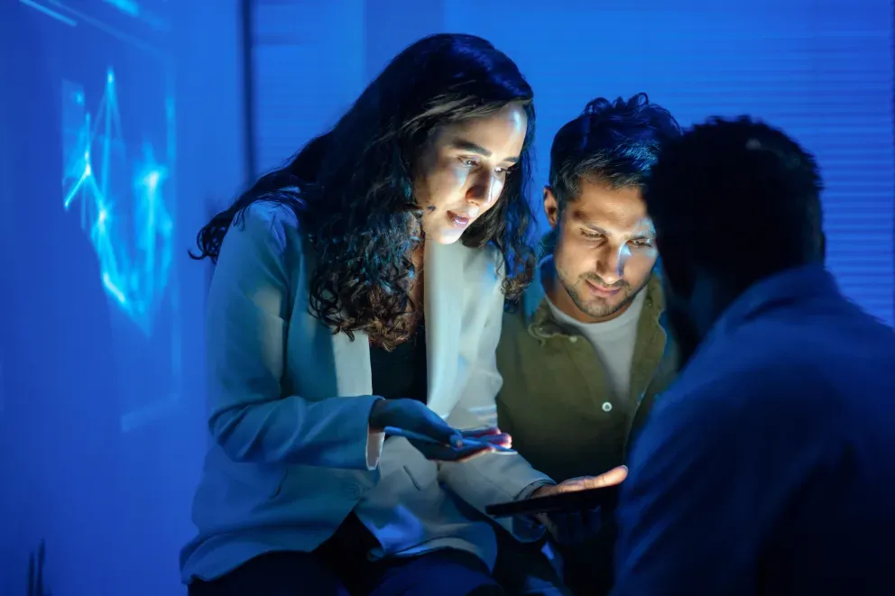 Three people gathered around a laptop computer in dim blue lighting, looking at the screen together. Three people gathered around a laptop computer in dim blue lighting, looking at the screen together.