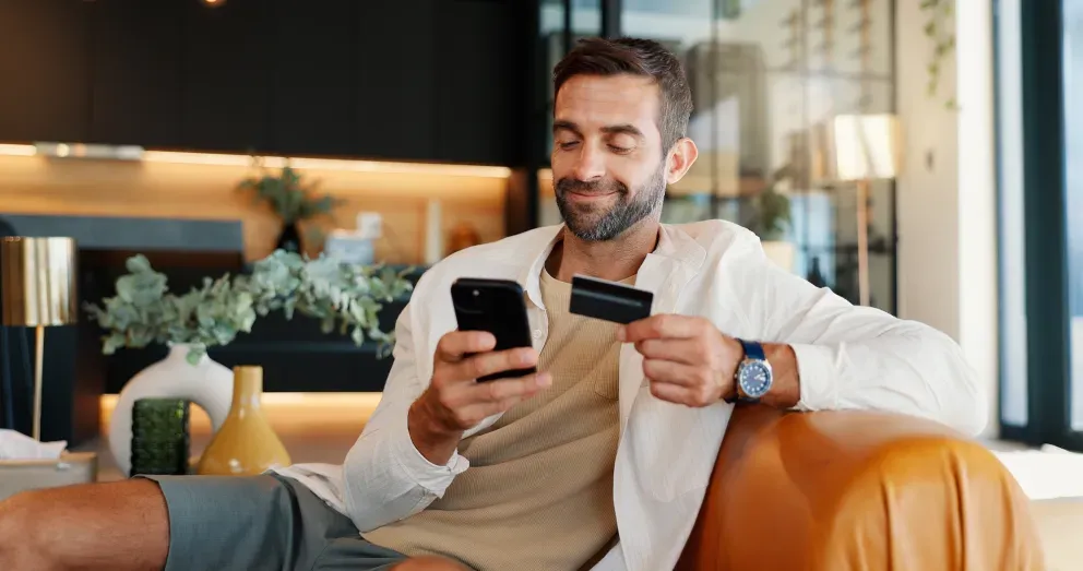 Lächelnder Mann, der auf einer Couch sitzt und ein Smartphone und eine Kreditkarte in der Hand hält, während er online einkauft.