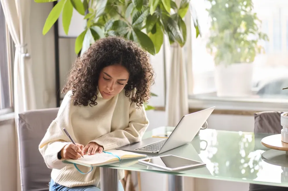 Junge Frau mit lockigem Haar schreibt Notizen an einem Glastisch mit einem aufgeklappten Laptop in der Nähe.