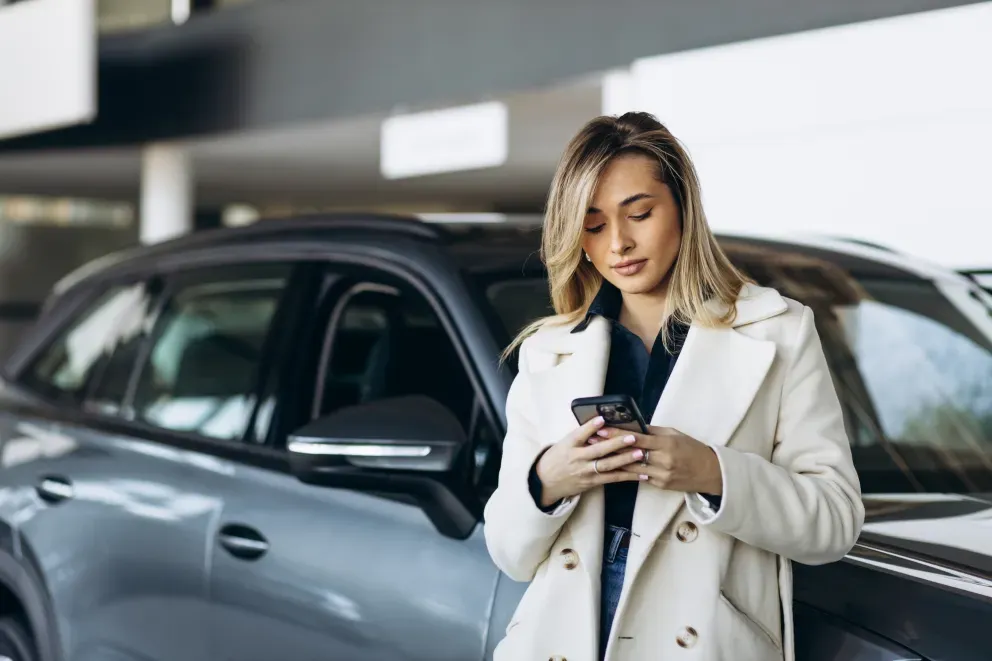 Frau in einem weißen Kittel schaut auf ihr Handy, während sie neben einem Auto in einem Autohaus steht.