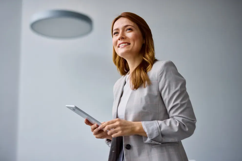 Lächelnde Geschäftsfrau in einem grauen Blazer, die ein Tablet in der Hand hält und selbstbewusst nach oben blickt. Lächelnde Geschäftsfrau in einem grauen Blazer, die ein Tablet in der Hand hält und selbstbewusst nach oben blickt.