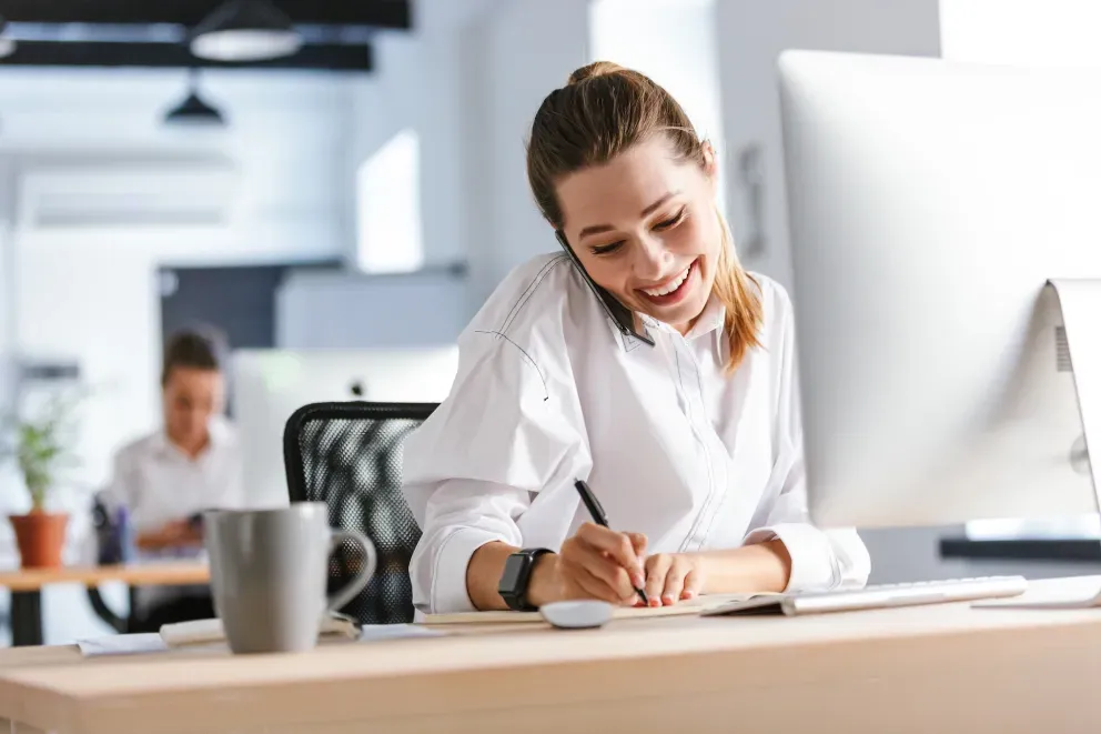 Smiling woman in a white shirt writing in a notebook at her desk in a bright office, with a gray mug nearby.