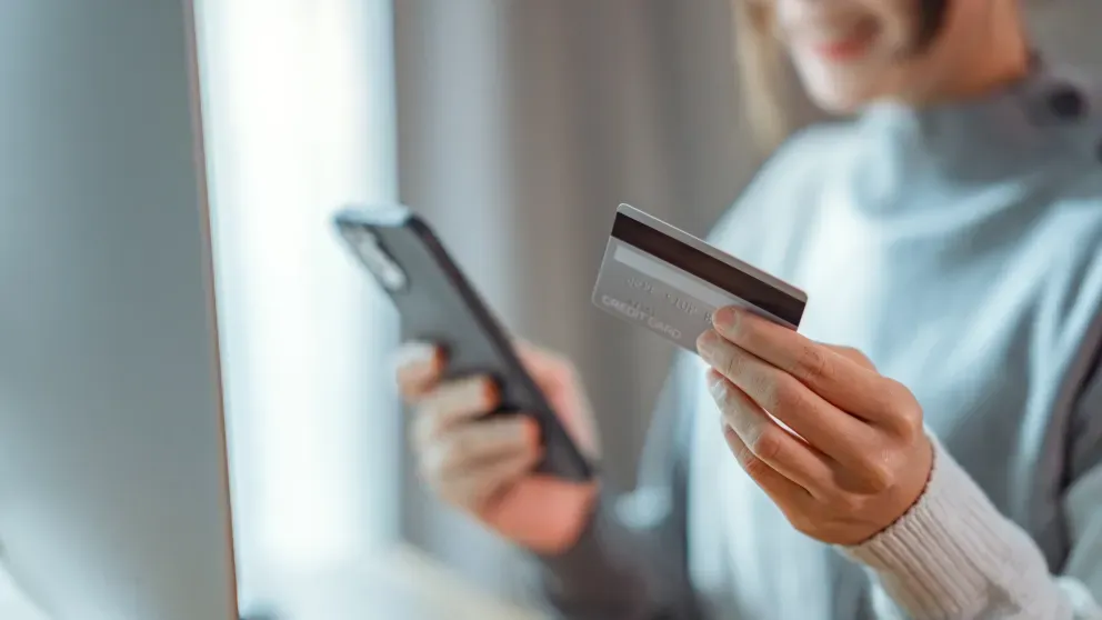 Person in a gray sweater holding a smartphone and a credit card, preparing to shop online by a window.