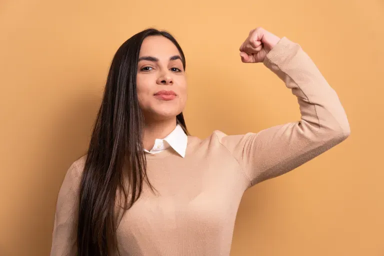 Junge Frau, die selbstbewusst ihren Bizeps vor einem beigen Hintergrund anspannt. Junge Frau, die selbstbewusst ihren Bizeps vor einem beigen Hintergrund anspannt.