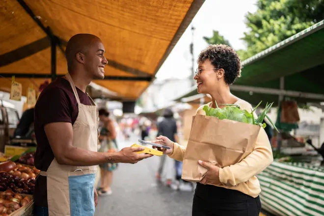 Smiling vendor in apron hands payment device to happy customer holding grocery bag at outdoor market. Smiling vendor in apron hands payment device to happy customer holding grocery bag at outdoor market.