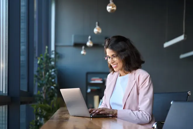 Woman in pink blazer smiling while working on a laptop at a modern office desk. Woman in pink blazer smiling while working on a laptop at a modern office desk.