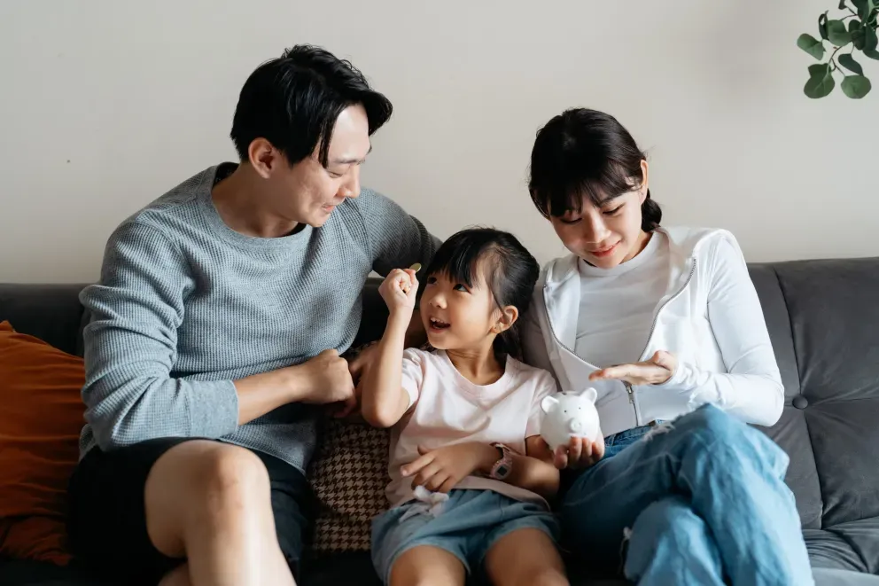 Familia feliz de tres sentados juntos en un sofá gris, padres con alcancía y un niño pequeño entre ellos