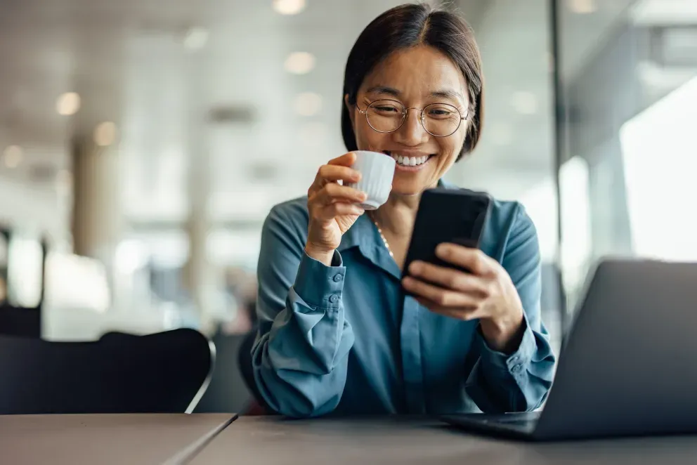 Joven sonriente con anteojos sosteniendo una taza de café y mirando un teléfono inteligente en la oficina.