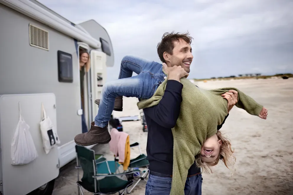 Hombre cargando a un niño, riendo junto a una casa rodante estacionada en una playa de arena.