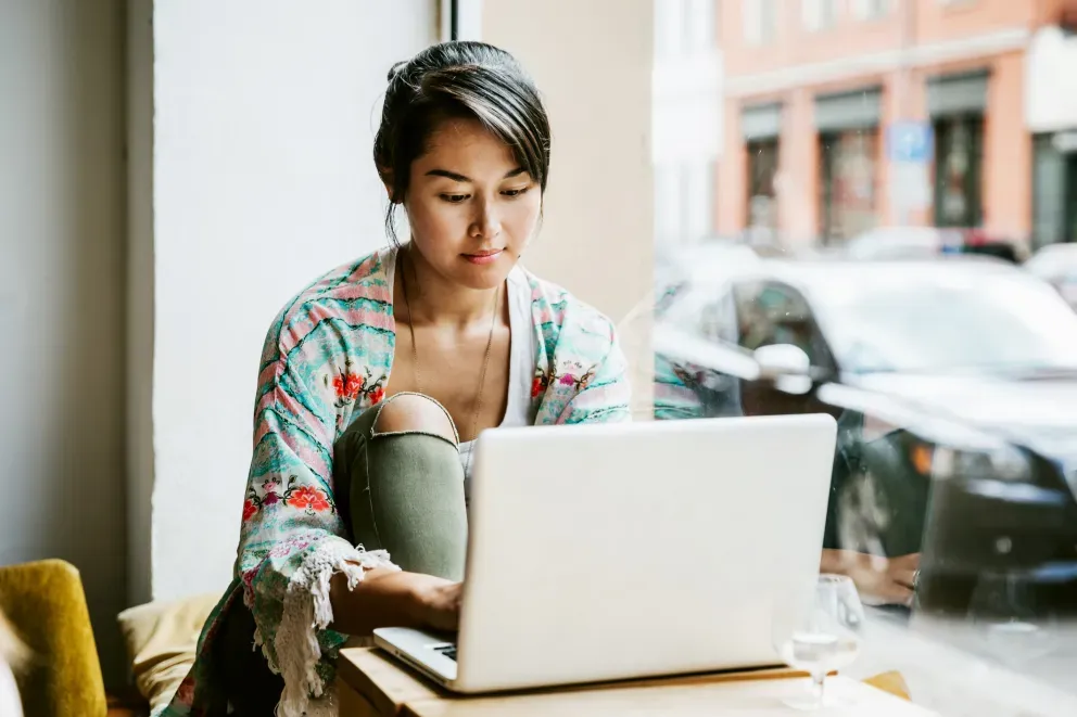 Young woman in floral top focused on laptop at a café window seat.
