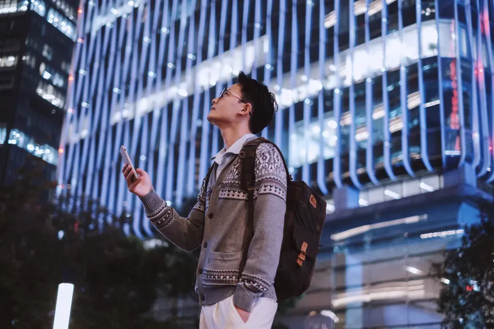 Joven mirando su teléfono por la noche, con los edificios iluminados de la ciudad detrás de él.