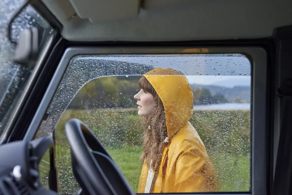 Mujer joven con un impermeable amarillo brillante sonriendo afuera de un vehículo en un día lluvioso.