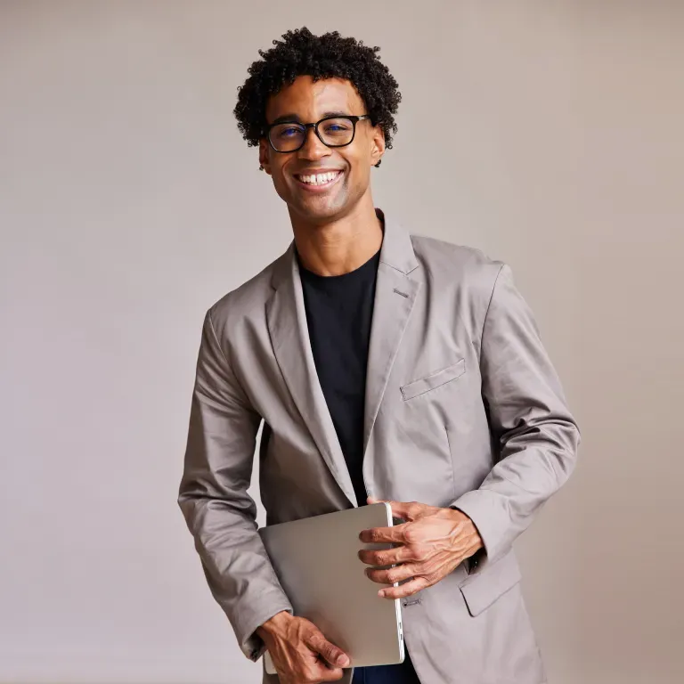 Young man with curly hair and glasses wearing a gray blazer, holding a laptop, smiling against a neutral background.