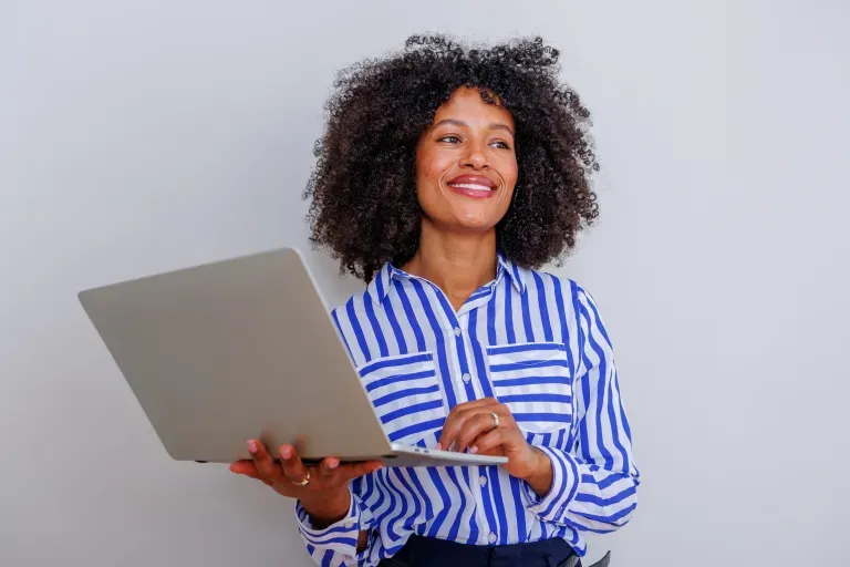 Smiling woman with curly hair in blue striped shirt holding laptop against gray background