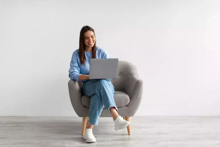 Mujer joven con atuendo azul sentada en un sillón gris, sonriendo mientras usa una computadora portátil.
