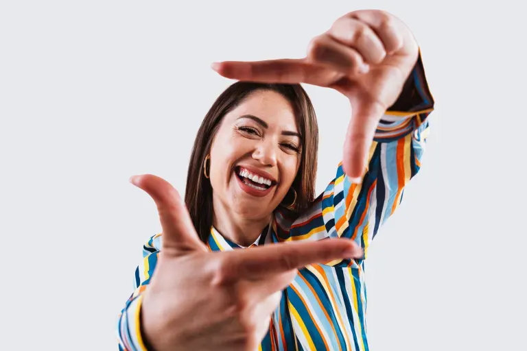 Mujer joven con camisa a rayas de colores que enmarca su rostro sonriente con las manos.