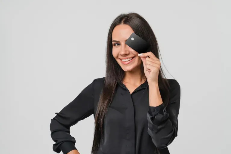 Mujer sonriente con blusa negra sosteniendo una pequeña tarjeta oscura cerca de su ojo sobre un fondo gris claro. Mujer sonriente con blusa negra sosteniendo una pequeña tarjeta oscura cerca de su ojo sobre un fondo gris claro.