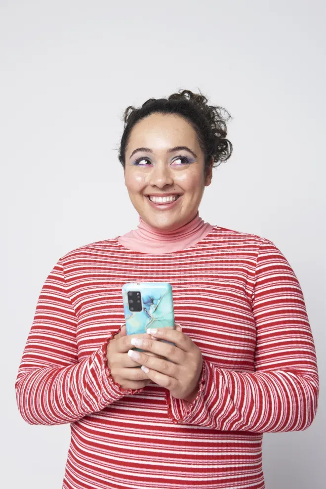 Smiling woman in red striped shirt holding a white smartphone against gray background