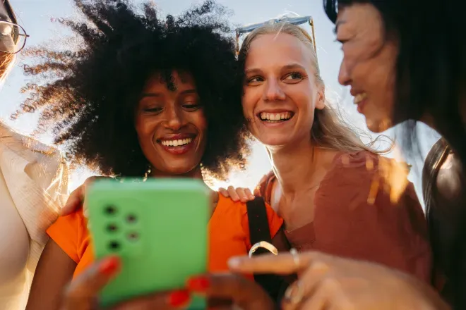 Tres mujeres jóvenes sonriendo y mirando juntas un teléfono inteligente verde al aire libre. Tres mujeres jóvenes sonriendo y mirando juntas un teléfono inteligente verde al aire libre.