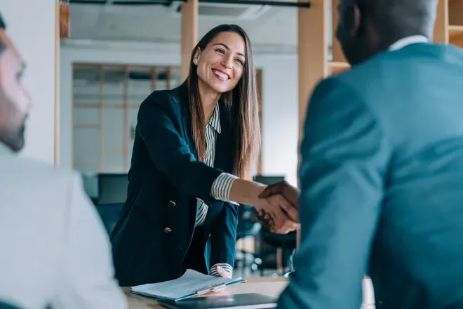 Mujer de negocios sonriente con un blazer negro estrechando la mano de un colega en una reunión profesional. Mujer de negocios sonriente con un blazer negro estrechando la mano de un colega en una reunión profesional.
