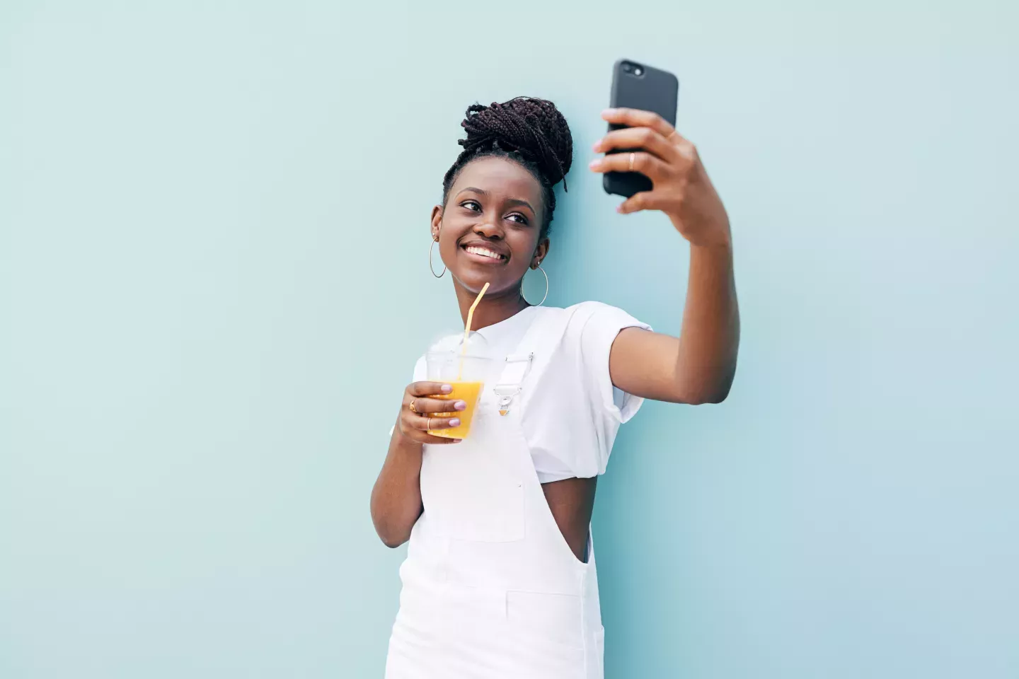 mujer joven sonriente apoyada en una pared azul claro sosteniendo jugo de naranja sosteniendo un teléfono celular