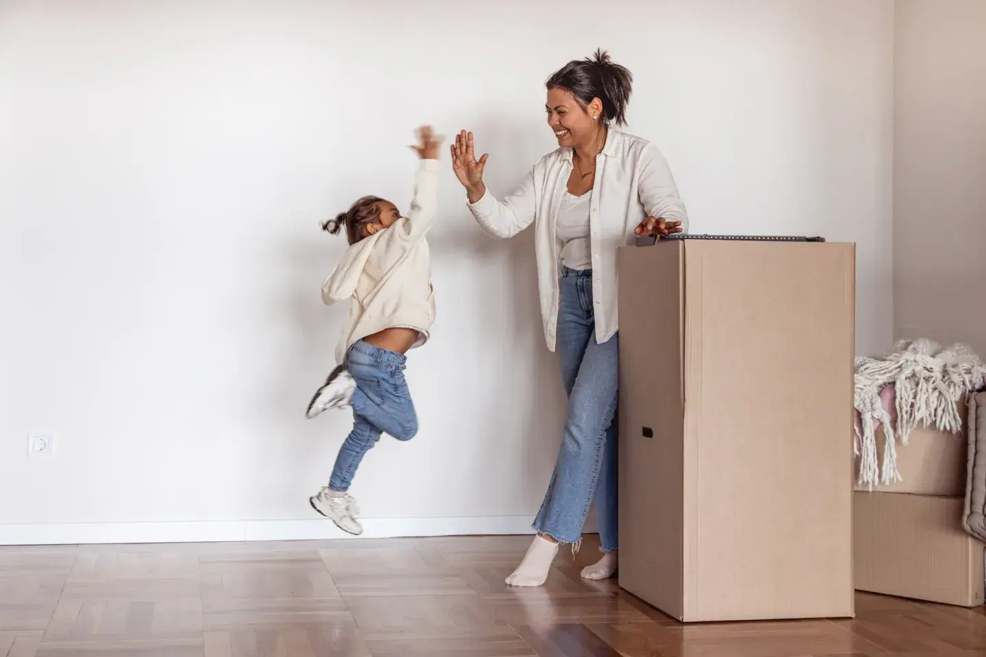 Mother and young daughter high-fiving joyfully next to a large cardboard moving box.