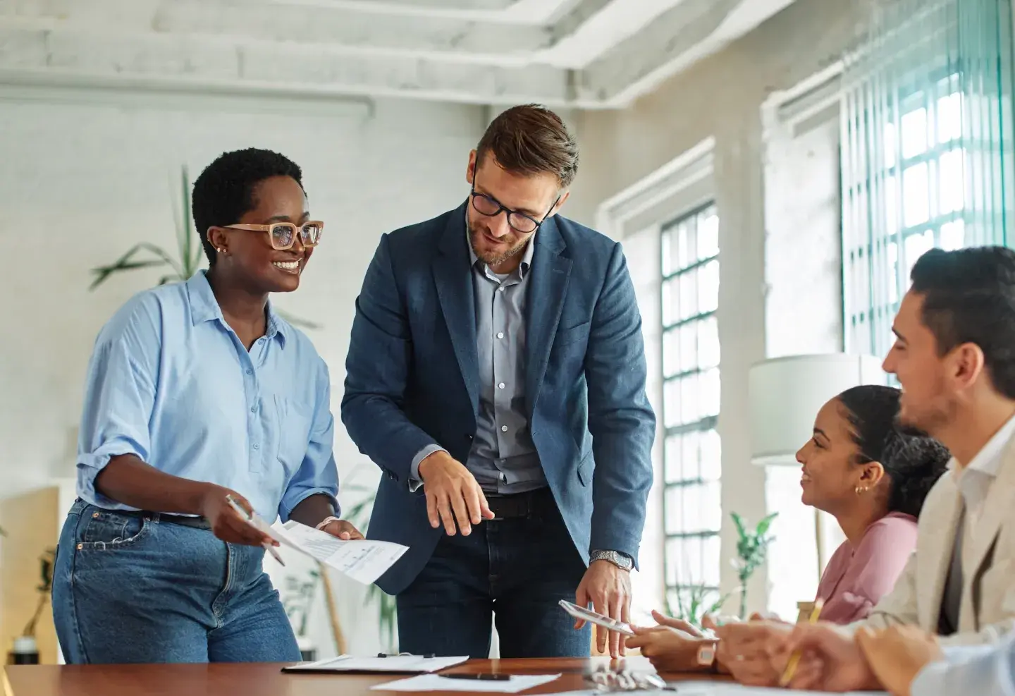 Two colleagues smiling and discussing work at a bright office table with coworkers nearby. Two colleagues smiling and discussing work at a bright office table with coworkers nearby.