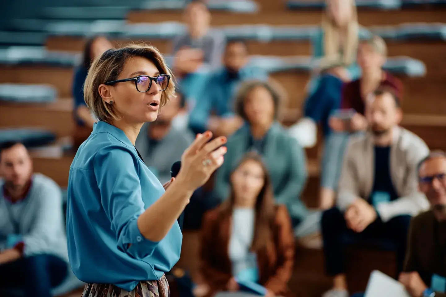 Woman in blue shirt with glasses speaking to a seated audience in a lecture hall. Woman in blue shirt with glasses speaking to a seated audience in a lecture hall.