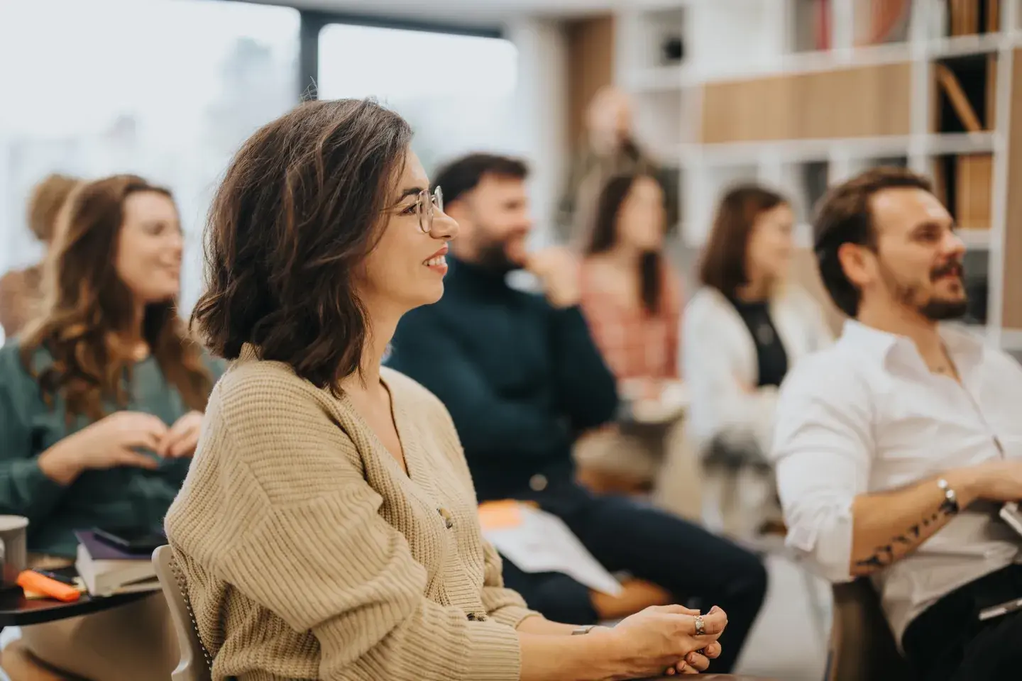Smiling woman with glasses seated among a group of people in a bright, casual meeting or workshop setting. Smiling woman with glasses seated among a group of people in a bright, casual meeting or workshop setting.
